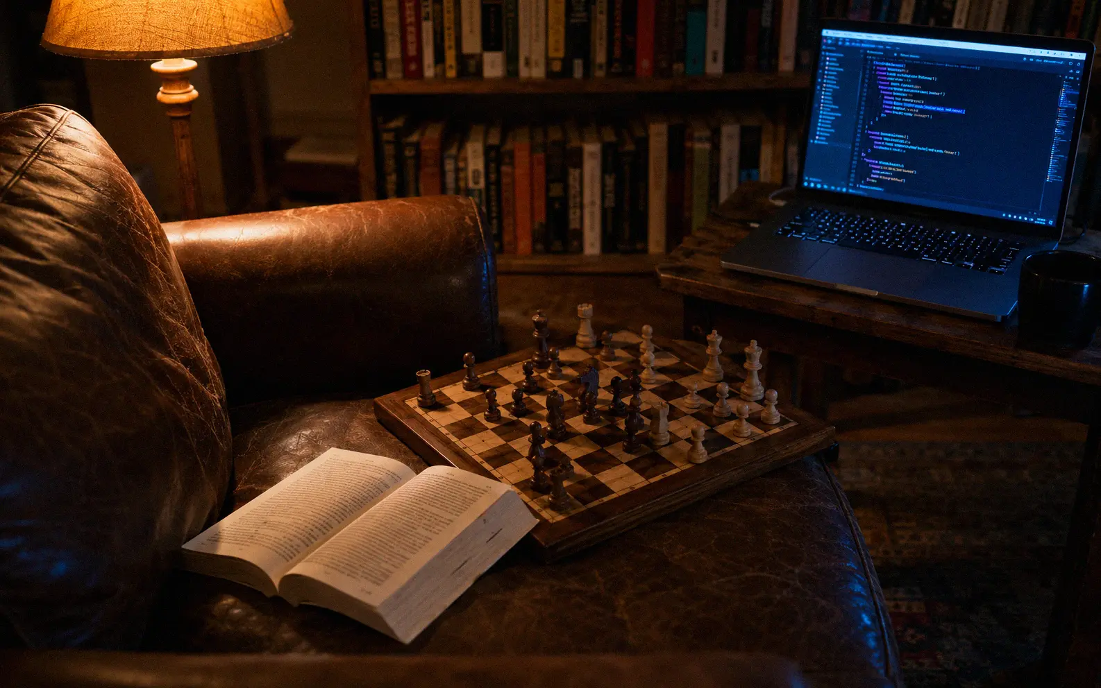 Open book on a leather chair next to a chess board mid-game and a laptop showing code